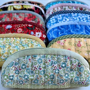K&ouml;nnte beinhalten: Eine Sammlung halbkreisf&ouml;rmiger, gesteppter Stofftaschen, die in Reihen angeordnet sind. Die Taschen weisen verschiedene Blumen- und geometrische Muster in den Farben Rot, Blau, Gelb, Gr&uuml;n und Schwarz auf, jeweils mit einem einfarbigen Stoffrand.