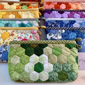 May include: A collection of colorful patchwork fabric pouches with hexagonal patterns. The pouches are arranged in a row, with the green pouch in the foreground. The pouches are all handmade and feature a variety of colors and patterns.