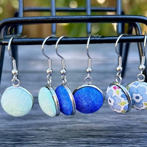 May include: A display of six fabric-covered button earrings with silver-colored hooks. The earrings are in various shades of blue and a floral pattern, displayed on a black metal stand. The earrings are round and have a fabric texture.