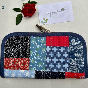 May include: A patchwork zippered pouch with a variety of fabric patterns in red, blue, and white. The pouch has a dark blue denim border and a metal zipper. A red rose and a business card with the text "Magnolia Handmade" are also visible.