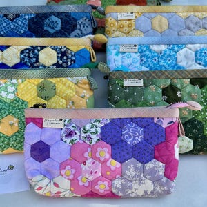 May include: A collection of handmade, quilted zipper pouches in various colors and patterns. The pouches feature a patchwork design with hexagonal fabric pieces. Each pouch has a different color scheme, including blue, yellow, green, and pink. The pouches are labeled with the brand name "Magnolia Handmade".