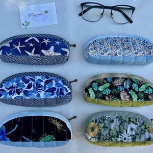 K&ouml;nnte beinhalten: Sechs halbkreisf&ouml;rmige Stoffetuis mit Rei&szlig;verschl&uuml;ssen, nummeriert von 50 bis 55, mit verschiedenen Mustern wie Seesternen, blauen Blumen, Libellen, kleinen Blumenprints, tropischen Bl&auml;ttern und Sonnenblumen, dazu eine Brille mit schwarzem Rahmen.