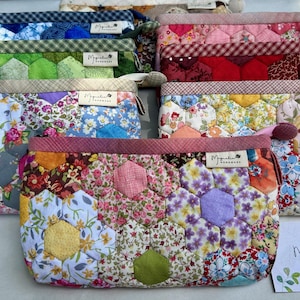 May include: A collection of rectangular zippered pouches made from quilted fabric featuring a hexagonal patchwork pattern in various floral and geometric prints. Each pouch has a small white fabric tag labeled Magnolia Handmade.