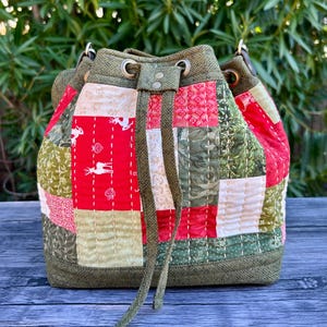May include: A patchwork bucket bag with a drawstring closure. The bag features a mix of red, green, and cream-colored fabric patches with white stitching. Some patches have deer and heart patterns. The top and base are a solid olive green.