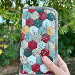 May include: A gray quilted wallet with a zipper closure and a chain. The wallet is decorated with a patchwork of hexagonal fabric pieces in shades of red, green, white, and gold. The fabric features a pattern of leaves and berries.