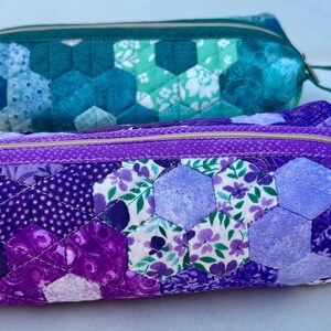 May include: Two quilted pencil cases. The bottom case is purple with a floral and polka dot pattern. The top case is teal with a hexagonal pattern. Both have zippers and wrist straps.