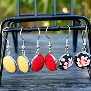 May include: Six pairs of dangle earrings with silver hooks are displayed on a black metal stand. The earrings feature fabric-covered circular pendants in yellow, red, and black with floral patterns. The earrings are a jewelry item.