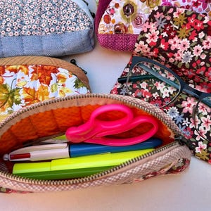 K&ouml;nnte beinhalten: Eine Sammlung von gesteppten Stoffetuis mit verschiedenen Blumen- und Herbstblattmustern. Ein offenes Etui enth&auml;lt eine leuchtend rosa Schere, einen mehrfarbigen Stift und einen gelben Textmarker. Eine Brille mit schwarzem Rahmen liegt auf einem Blumenetui.
