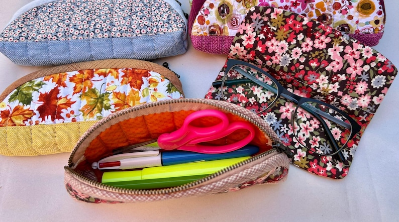 K&ouml;nnte beinhalten: Eine Sammlung von gesteppten Stoffetuis mit verschiedenen Blumen- und Herbstblattmustern. Ein offenes Etui enth&auml;lt eine leuchtend rosa Schere, einen mehrfarbigen Stift und einen gelben Textmarker. Eine Brille mit schwarzem Rahmen liegt auf einem Blumenetui.