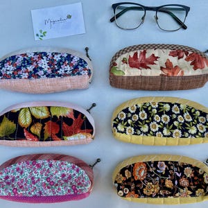 K&ouml;nnte beinhalten: Sechs halbkreisf&ouml;rmige Stoffetuis mit Rei&szlig;verschluss, nummeriert von 14 bis 19, mit verschiedenen Blumen- und Blattmustern in Farben wie Rosa, Gelb, Braun und Schwarz, dazu eine schwarz umrandete Brille und eine Karte mit Magnolia Handmade.