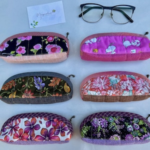 K&ouml;nnte beinhalten: Sechs gesteppte Stoffetuis mit Rei&szlig;verschluss, nummeriert von 20 bis 25. Die Etuis zeigen verschiedene Blumen- und Cartoon-Motive in Rosa, Lila, Schwarz und Gr&uuml;n. Eine schwarze Brille und eine Karte mit der Aufschrift Magnolia Handmade liegen bei.