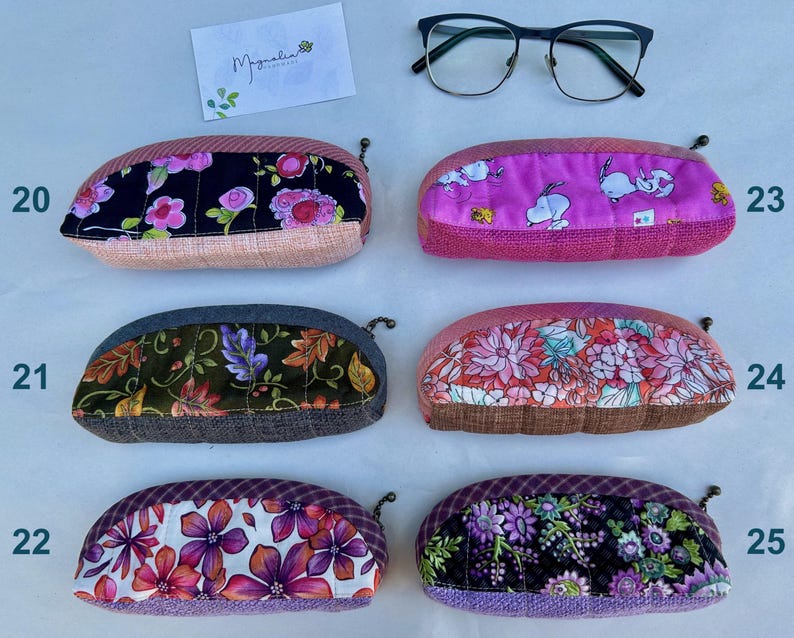 K&ouml;nnte beinhalten: Sechs gesteppte Stoffetuis mit Rei&szlig;verschluss, nummeriert von 20 bis 25. Die Etuis zeigen verschiedene Blumen- und Cartoon-Motive in Rosa, Lila, Schwarz und Gr&uuml;n. Eine schwarze Brille und eine Karte mit der Aufschrift Magnolia Handmade liegen bei.