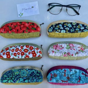 K&ouml;nnte beinhalten: Sechs gesteppte Brillenetuis aus Stoff mit verschiedenen Mustern wie Erdbeeren, Blumen und Libellen, nummeriert von 56 bis 61. Eine Brille mit schwarzem Rahmen und eine wei&szlig;e Visitenkarte mit dem Text Magnolia befinden sich &uuml;ber den Etuis.