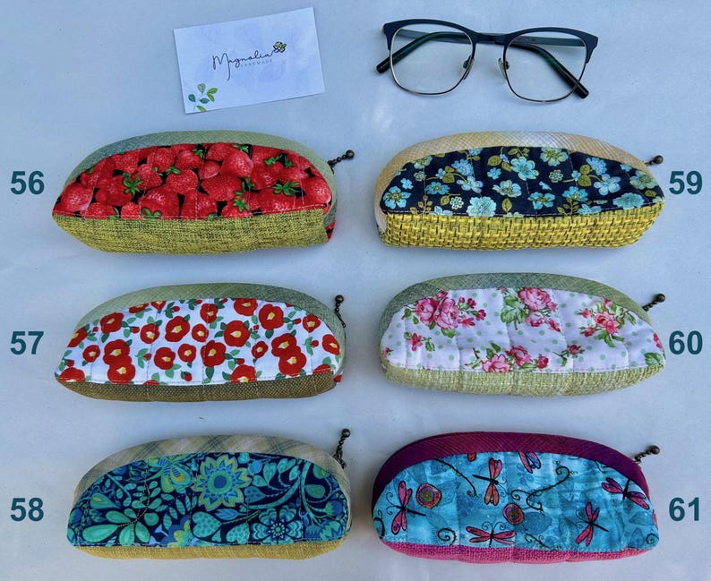 K&ouml;nnte beinhalten: Sechs gesteppte Brillenetuis aus Stoff mit verschiedenen Mustern wie Erdbeeren, Blumen und Libellen, nummeriert von 56 bis 61. Eine Brille mit schwarzem Rahmen und eine wei&szlig;e Visitenkarte mit dem Text Magnolia befinden sich &uuml;ber den Etuis.