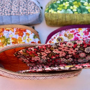 K&ouml;nnte beinhalten: Eine Sammlung von f&uuml;nf halbkreisf&ouml;rmigen, gesteppten Stofftaschen mit verschiedenen Mustern, darunter kleine Blumenprints, gr&uuml;ne Kleebl&auml;tter, Herbstbl&auml;tter und leuchtend rosa Bl&uuml;ten, jeweils mit einem Metallrei&szlig;verschluss und gestepptem Innenfutter.