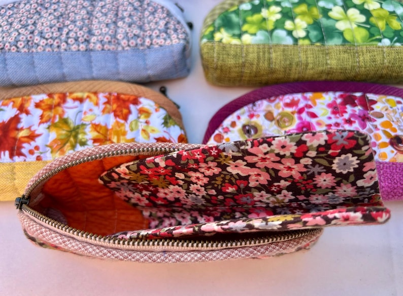 K&ouml;nnte beinhalten: Eine Sammlung von f&uuml;nf halbkreisf&ouml;rmigen, gesteppten Stofftaschen mit verschiedenen Mustern, darunter kleine Blumenprints, gr&uuml;ne Kleebl&auml;tter, Herbstbl&auml;tter und leuchtend rosa Bl&uuml;ten, jeweils mit einem Metallrei&szlig;verschluss und gestepptem Innenfutter.