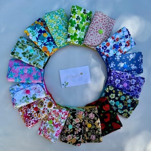 May include: A collection of colorful fabric wallets arranged in a circle. Each wallet features a unique floral pattern and a button closure. The wallets are in various colors, including blue, green, pink, and red. A small card with the text "Magnolia" is in the center.