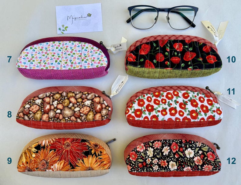 K&ouml;nnte beinhalten: Sechs halbkreisf&ouml;rmige, gesteppte Etuis mit Rei&szlig;verschl&uuml;ssen, nummeriert von 7 bis 12. Die Designs umfassen bunte Herzen, rote Mohnblumen auf Schwarz, braune Steine, rote Blumen auf Wei&szlig;, orangefarbene Sonnenblumen und kleine Blumenmuster.