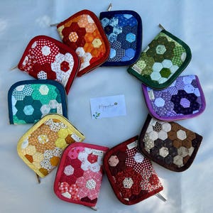 May include: A collection of ten zippered pouches arranged in a circle, each featuring a quilted hexagonal patchwork pattern in various colors including red, orange, blue, green, purple, yellow, pink, and brown, with a central card reading Magnolia.