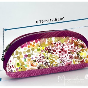 K&ouml;nnte beinhalten: Ein Rei&szlig;verschlussetui mit Blumenmuster in Rosa-, Gelb- und Gr&uuml;nt&ouml;nen und einem einfarbigen violetten Rand. Das Etui misst 17,5 cm in der L&auml;nge, 8 cm in der H&ouml;he und 3,5 cm in der Tiefe. Der Text Magnolia Handmade ist sichtbar.