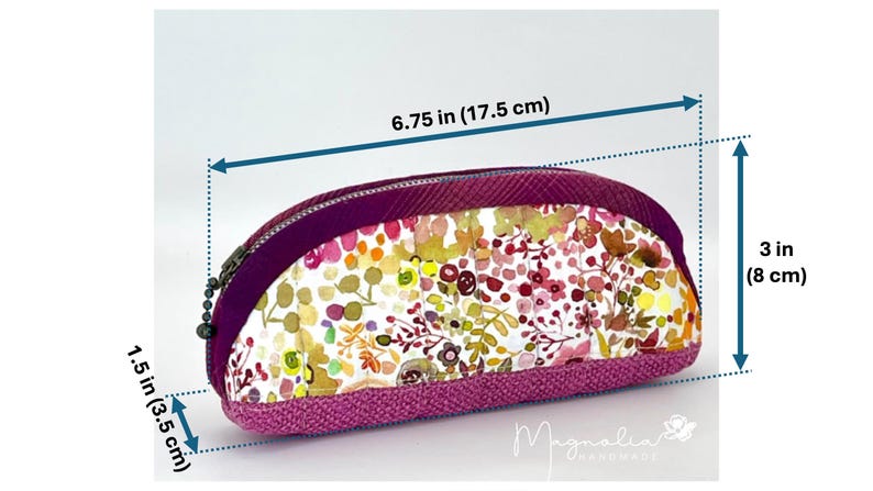 K&ouml;nnte beinhalten: Ein Rei&szlig;verschlussetui mit Blumenmuster in Rosa-, Gelb- und Gr&uuml;nt&ouml;nen und einem einfarbigen violetten Rand. Das Etui misst 17,5 cm in der L&auml;nge, 8 cm in der H&ouml;he und 3,5 cm in der Tiefe. Der Text Magnolia Handmade ist sichtbar.