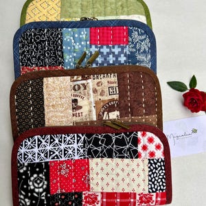 May include: A stack of four handmade quilted zipper pouches in various colors and patterns. The pouches feature patchwork designs with fabrics in green, blue, brown, and red. Each pouch has a contrasting zipper and a quilted texture.