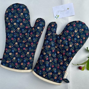 May include: A pair of quilted oven mitts featuring a dark blue fabric base with a repeating pattern of small pink, orange, and blue flowers with green leaves. Each mitt has a cream-colored trim at the opening and a small fabric tag on the side.