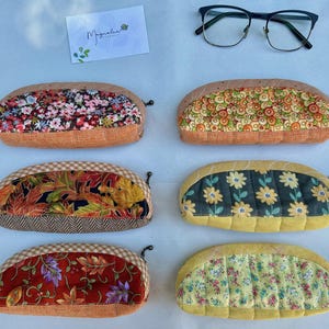 K&ouml;nnte beinhalten: Sechs halbkreisf&ouml;rmige Stoffetuis mit den Nummern 32 bis 37, die verschiedene Blumen-, Blatt- und geometrische Muster aufweisen. Jedes Etui hat einen Rei&szlig;verschluss und eine strukturierte Basis. Eine schwarz umrandete Brille und eine wei&szlig;e Karte mit dem Text Magnolia sind enthalten.