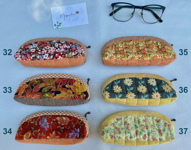 K&ouml;nnte beinhalten: Sechs halbkreisf&ouml;rmige Stoffetuis mit den Nummern 32 bis 37, die verschiedene Blumen-, Blatt- und geometrische Muster aufweisen. Jedes Etui hat einen Rei&szlig;verschluss und eine strukturierte Basis. Eine schwarz umrandete Brille und eine wei&szlig;e Karte mit dem Text Magnolia sind enthalten.
