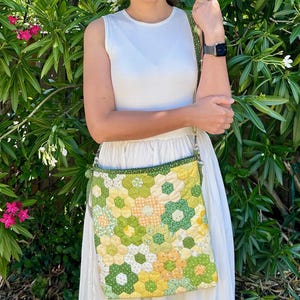 May include: A quilted crossbody bag with a floral pattern in shades of green, yellow, and white. The bag features a green strap and trim, and a silver chain. The bag is approximately 10 inches wide and 10 inches tall.
