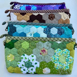 May include: A stack of handmade fabric pouches with hexagonal patchwork designs. The pouches feature various colors, including green, blue, brown, and purple. Each pouch has a zipper closure and a small loop on the side.