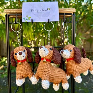 May include: Three crochet dog keychains. Each dog has brown ears, a brown tail, and a tan body with white paws. They wear red collars with a small gold bell. The keychains hang from a black metal display stand.