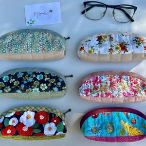 K&ouml;nnte beinhalten: Sechs halbkreisf&ouml;rmige Rei&szlig;verschlusstaschen mit gesteppten Blumenstoffbahnen und strukturierten Unterteilen, nummeriert von 26 bis 31. Eine wei&szlig;e Karte mit dem Text Magnolia Handmade und eine schwarz umrandete Brille liegen dar&uuml;ber.