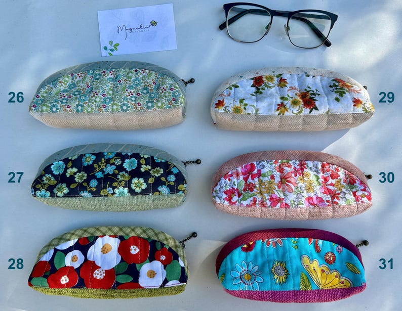 K&ouml;nnte beinhalten: Sechs halbkreisf&ouml;rmige Rei&szlig;verschlusstaschen mit gesteppten Blumenstoffbahnen und strukturierten Unterteilen, nummeriert von 26 bis 31. Eine wei&szlig;e Karte mit dem Text Magnolia Handmade und eine schwarz umrandete Brille liegen dar&uuml;ber.