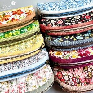 K&ouml;nnte beinhalten: Eine Sammlung von zehn halbkreisf&ouml;rmigen, gesteppten Taschen mit verschiedenen Blumen-, Blatt- und anderen Mustern. Jede Tasche hat einen Metallrei&szlig;verschluss und eine strukturierte Stoffkante. Der Text Magnolia Handmade ist oben links sichtbar.