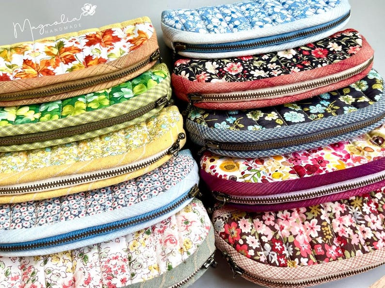 K&ouml;nnte beinhalten: Eine Sammlung von zehn halbkreisf&ouml;rmigen, gesteppten Taschen mit verschiedenen Blumen-, Blatt- und anderen Mustern. Jede Tasche hat einen Metallrei&szlig;verschluss und eine strukturierte Stoffkante. Der Text Magnolia Handmade ist oben links sichtbar.