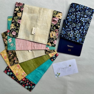 May include: A collection of fabric passport covers and book covers in various floral patterns and colors. One cover is open, displaying a blue passport. The covers feature a mix of floral prints, including pink, yellow, and blue flowers. A business card with the text "Mygnalia Handmade" is also present.