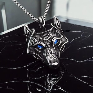 May include: A silver wolf head pendant necklace with blue gemstone eyes. The pendant has detailed engravings and hangs from a matching chain. The necklace is shown on a black surface.