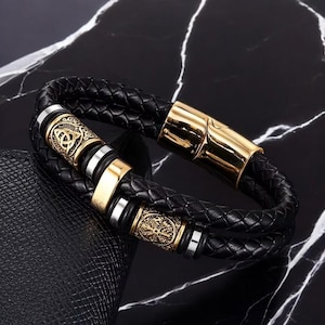 May include: Black braided leather bracelet with gold-tone and silver-tone accents. The bracelet features decorative gold-tone beads with intricate designs and a gold-tone clasp. The bracelet is approximately 8 inches (20 cm) in length.