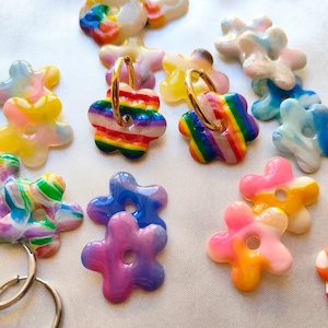 May include: A collection of colorful, flower-shaped earrings and charms. The earrings feature gold hoops and rainbow-striped flower pendants. Other charms display a variety of colors, including pink, blue, yellow, and green, with a glossy finish. The items are arranged on a white surface.