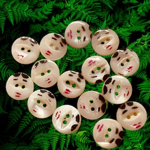 May include: A collection of round, cream-colored buttons, each featuring a hand-painted face with dark brown hair, eyebrows, and red lips. Each button has two central holes for attachment, with varying facial expressions and hair styles depicted.