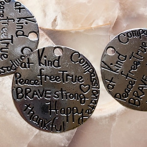 May include: Silver charm with a circular design featuring the words "Peace free True", "at Kind", "Brave Strong", "Happy", "Thankful", and "Compassionate".