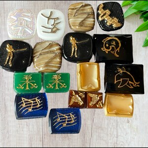 Pode incluir: Uma coleção de cabochões decorativos de vidro em várias formas e cores. Os desenhos incluem notas musicais, figuras estilizadas e padrões abstratos em ouro, branco, castanho, verde e azul. Os cabochões estão sobre um fundo claro com grão de madeira.