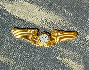 Puede incluir: Un pin de ala de piloto de color dorado con una pequeña esfera de reloj en el centro. El reloj tiene una esfera blanca con números romanos y manecillas negras. Las alas están detalladas con líneas en forma de pluma.