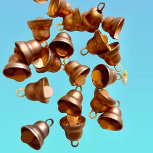 May include: A collection of small, matte copper-colored metal bells scattered across the frame. Each bell features a rounded body, a flared opening at the bottom, and a small circular loop at the top for attachment, with a clapper visible inside.