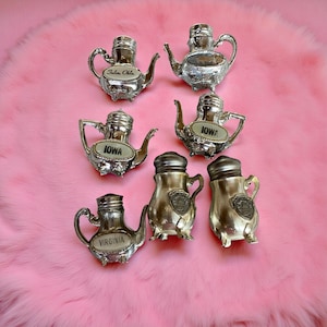May include: A collection of vintage silver-toned metal teapot and creamer salt and pepper shakers. Some shakers have the names of states, including Iowa, Oklahoma, and Virginia. The items are arranged on a pink, fluffy surface.
