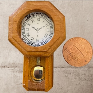 Puede incluir: Un reloj de madera en miniatura con una esfera blanca y números romanos negros. El reloj tiene un péndulo y es del tamaño de una moneda de un centavo de euro.