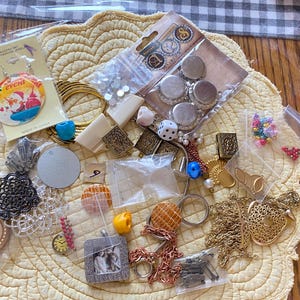 May include: A collection of jewelry making supplies including beads, charms, chains, and findings. The supplies are arranged on a white crocheted doily. The image also includes a small bag of rhinestones, a bag of small silver metal caps, and a bag of small colorful beads.