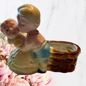 May include: A vintage ceramic planter featuring two figures in a loving embrace. The figures are painted with soft colors, including pink, blue, and yellow, with gold accents. The planter has a brown, textured exterior.