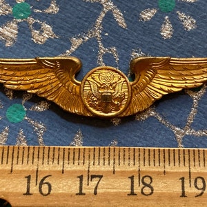 Puede incluir: Unas alas de piloto militar doradas con un emblema redondo dorado en el centro. El emblema presenta un &aacute;guila calva con un escudo en el pecho. Las alas miden aproximadamente 4,4 cm de ancho.